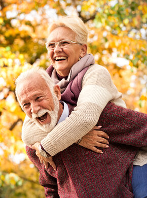 Ein älterer Mann mit rotem Pullover trägt eine ältere Frau mit weißem Pullover auf dem Rücken. Beide lachen. Im Hintergrund sind Bäume zu sehen.